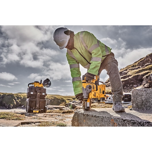 Worker uses DEWALT hammer drill on concrete outdoors, wearing safety gear. DEWALT tools visible in the background.