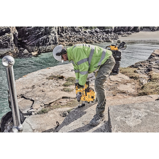 Worker using a DEWALT rotary hammer drill on a cracked concrete surface outdoors near water and rocks.
