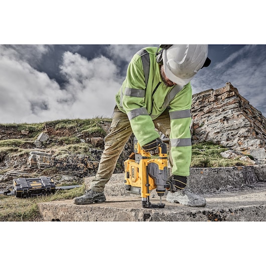 Worker using DEWALT rotary hammer drill on outdoor concrete, wearing safety gear and high-visibility clothing.