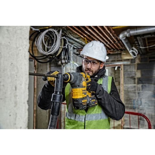 Worker using DEWALT DCH323NT rotary hammer drill on a concrete wall at a construction site, wearing safety gear.