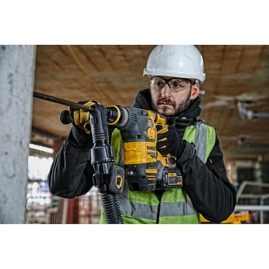 Worker uses DEWALT rotary hammer drill DCH323NT on construction site, wearing safety gear and hi-vis vest.