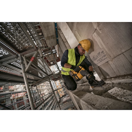 Worker uses DEWALT power tool on concrete, wearing safety gear and working near scaffolding at construction site.