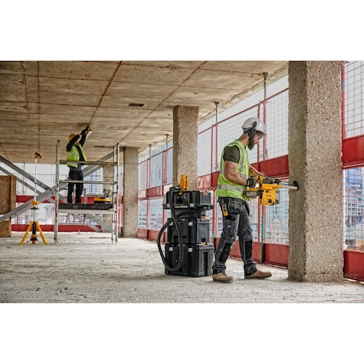 Two construction workers are operating DEWALT power tools at a building site. One worker is drilling into a concrete pillar, wearing a hard hat, safety vest, and protective gear, while the other worker is standing on scaffolding measuring the ceiling. DEWALT toolboxes and equipment are visible nearby.