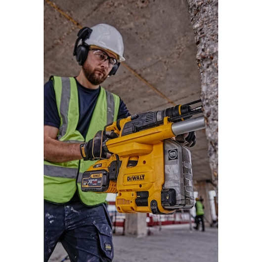 Worker holding a DEWALT DCH334X2 tool at a construction site, wearing safety gear and a green vest.