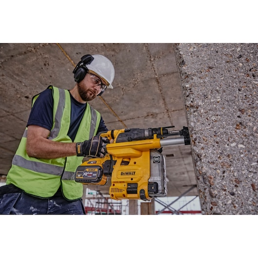A person wearing safety gear and a high-visibility vest operates a yellow DEWALT DCH334X2 rotary hammer drill against a concrete wall at a construction site.