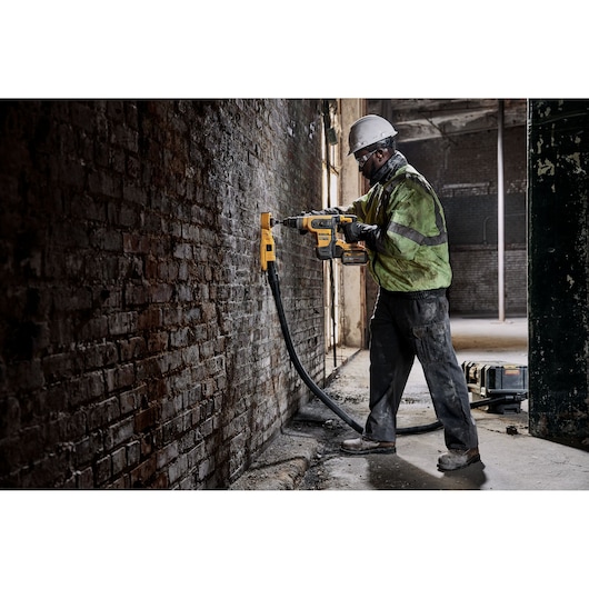 Worker uses DEWALT rotary hammer drill with vacuum on a brick wall in an industrial setting.