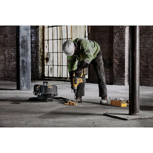 Worker drills concrete floor with DEWALT tool. Safety gear and accessories visible in industrial setting.