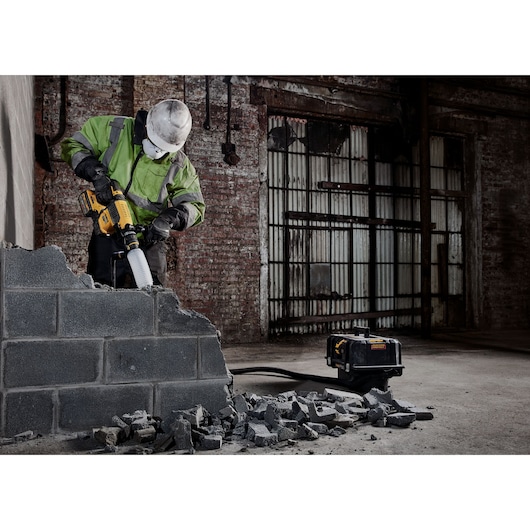 Worker uses DEWALT demolition tool on concrete wall, debris on ground, industrial background.