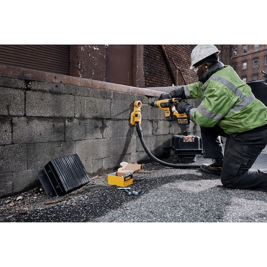 Worker drills a brick wall outdoors using a DEWALT tool, with parts and a case nearby.