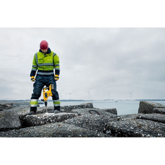 A person wearing high-visibility workwear and a red helmet is using a yellow DEWALT rotary hammer drill (SKU: DCH481N) to drill into concrete at an outdoor construction site by the water, with wind turbines visible in the background.