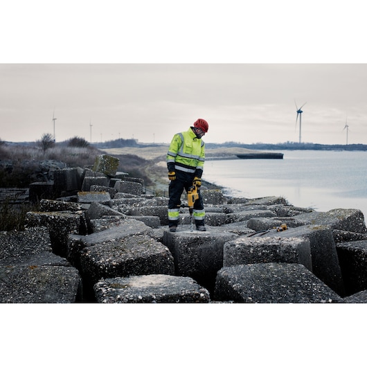 Worker in safety gear uses DEWALT tool on large concrete blocks near water with wind turbines in background.