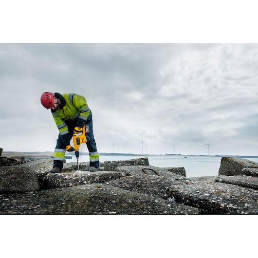Worker using yellow DEWALT tool outdoors on concrete, wearing safety gear. Wind turbines in background.