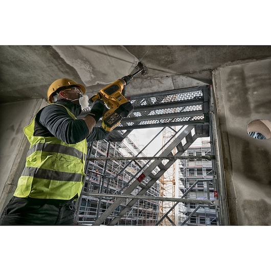 Worker uses DEWALT rotary hammer drill on concrete ceiling at a construction site with scaffolding.
