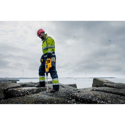 Worker uses a DEWALT demolition hammer on concrete outdoors, wearing helmet and safety gear.