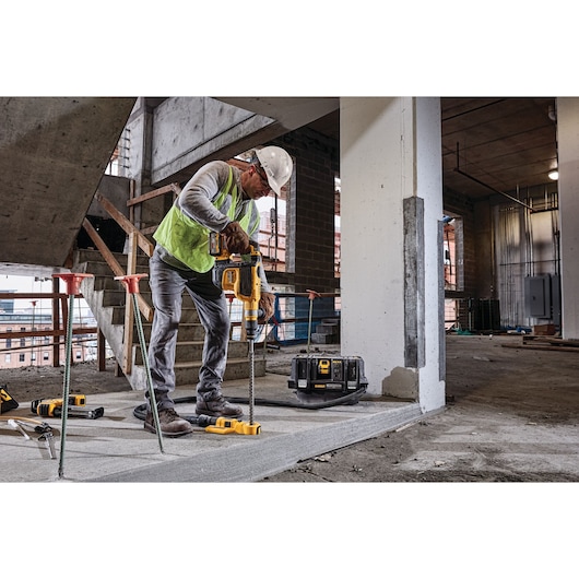 Worker using DEWALT rotary hammer drill on concrete at a construction site with safety gear and DEWALT toolbox.