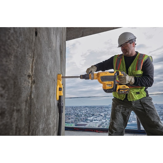 Worker uses DEWALT rotary hammer drill on a concrete wall at a construction site overlooking a cityscape.
