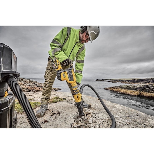 Person uses DEWALT rotary hammer drill to break concrete on a rocky shore with safety gear and vacuum hose.