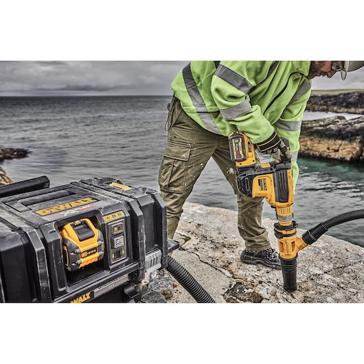DEWALT rotary hammer drilling concrete outdoors near water, with a worker and equipment case visible.