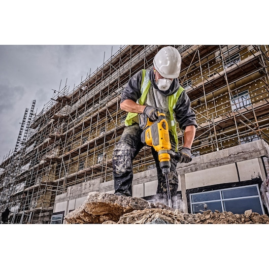 A construction worker using a yellow DEWALT rotary hammer drill on a rocky surface at a building site, with scaffolding and a partially built structure in the background. The worker is wearing safety gear, including a helmet, gloves, and a high-visibility vest.