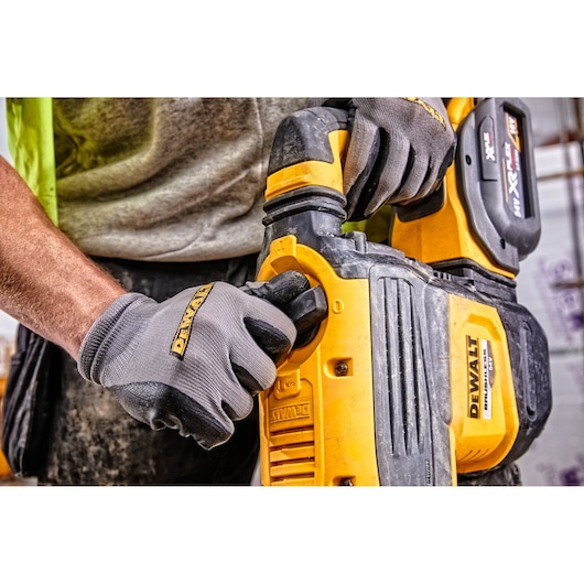 Close-up of a person wearing DEWALT gloves, operating a yellow DEWALT cordless power tool, likely a rotary hammer, with visible DEWALT branding and battery pack.