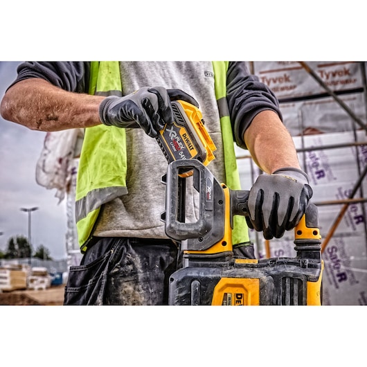 A person wearing a high visibility vest and work gloves is holding a DEWALT power tool, inserting a battery pack. Construction materials and scaffolding can be seen in the background.