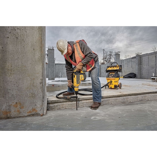 A construction worker wearing a hard hat and safety vest operates a DEWALT rotary hammer drill to drill into a concrete floor at an industrial construction site. A DEWALT dust extractor is visible nearby, connected by a hose to the drill.