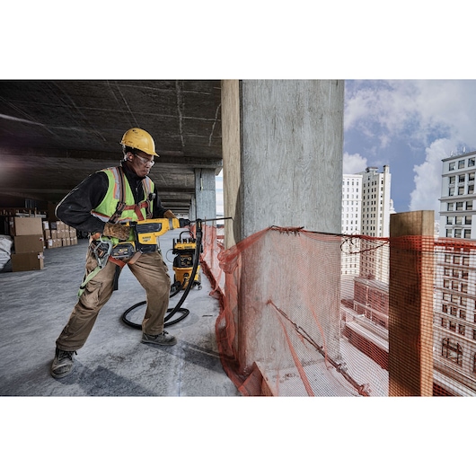 A construction worker wearing safety gear is using a DEWALT rotary hammer drill (model DCH733X2) on a concrete pillar at an unfinished building site, with safety netting along the edge and city buildings visible in the background.