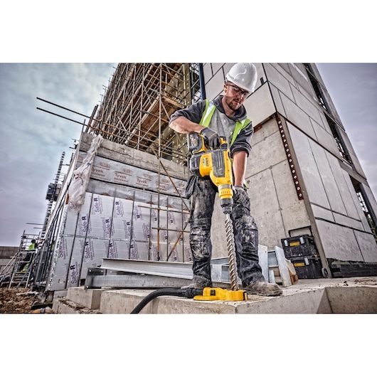 A construction worker wearing a hard hat and safety vest is using a yellow DEWALT rotary hammer drill (DCH733X2) on a concrete surface at an outdoor construction site, with scaffolding and building materials visible in the background.