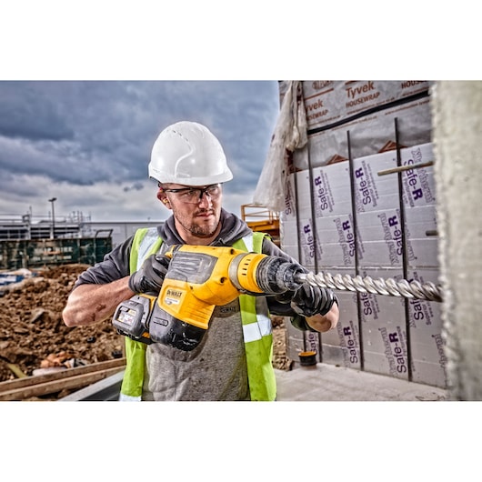 A construction worker wearing a white hard hat and safety vest operates a yellow DEWALT DCH733X2 rotary hammer drill with a large drill bit at a building site. The background shows construction materials and partially built structures.
