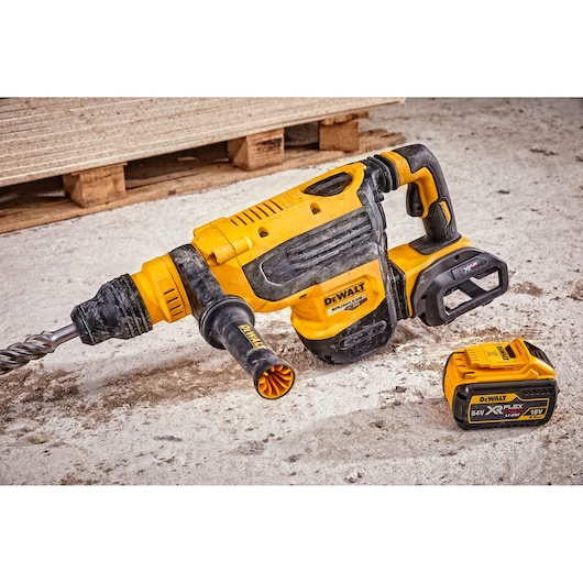 A DEWALT branded cordless rotary hammer drill with an attached handle, shown on a construction site floor. Next to it is a DEWALT XR FLEXVOLT 54V/18V lithium-ion battery.