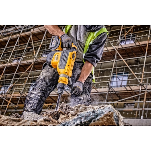 A construction worker uses a yellow DEWALT cordless rotary hammer drill (model DCH733X2) to break concrete at a construction site, with scaffolding and a building visible in the background.