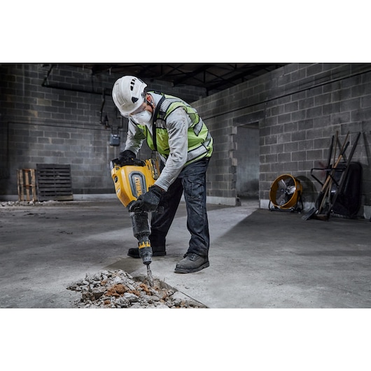 Worker uses a DEWALT demolition hammer to break concrete on a construction site, wearing safety gear.