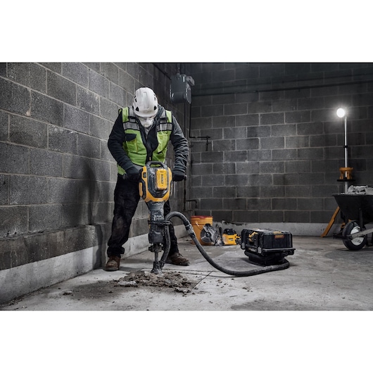 A construction worker in safety gear operates a DEWALT electric jackhammer to break up concrete on a worksite. The tool is connected to a vacuum system and there is construction equipment in the background.