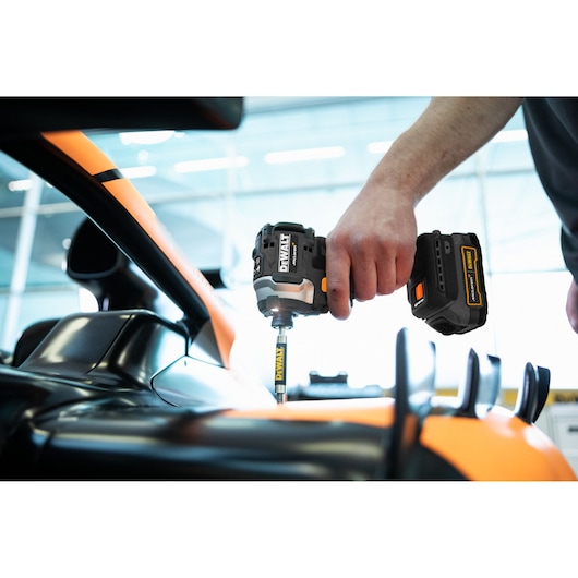 A person using a DEWALT cordless impact driver to work on a black and orange vehicle, possibly performing maintenance or assembly in a well-lit indoor environment.