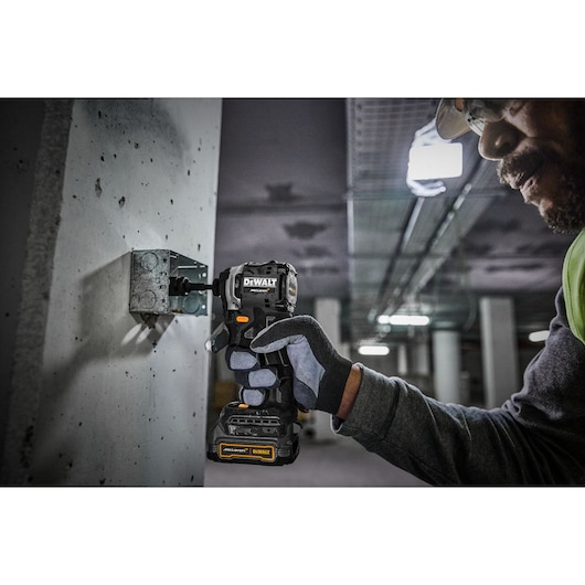 A person wearing safety gloves uses a DEWALT cordless power drill to secure a metal fixture to a concrete wall in an industrial setting.