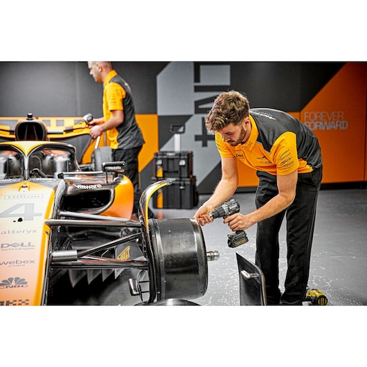 A person in a yellow and black uniform uses a DEWALT power tool to work on the wheel assembly of an orange Formula 1 race car. Another person in the background also wears a similar uniform. The setting appears to be a garage or workshop with toolboxes and equipment visible.