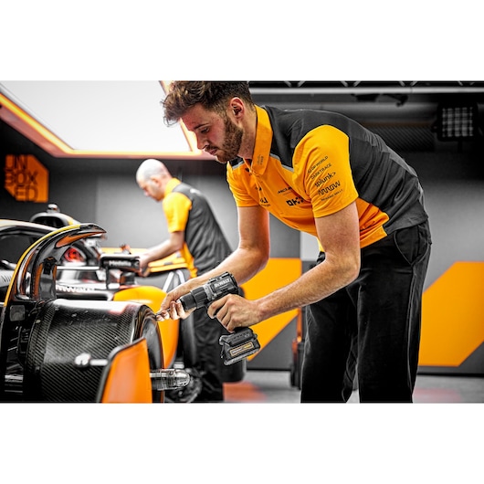 A person in a black and orange uniform uses a DEWALT cordless drill on a race car in a garage or workshop, with another person working in the background.