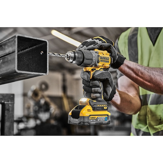 Person drills metal with a DEWALT cordless drill in a workshop. Wearing gloves and safety vest.