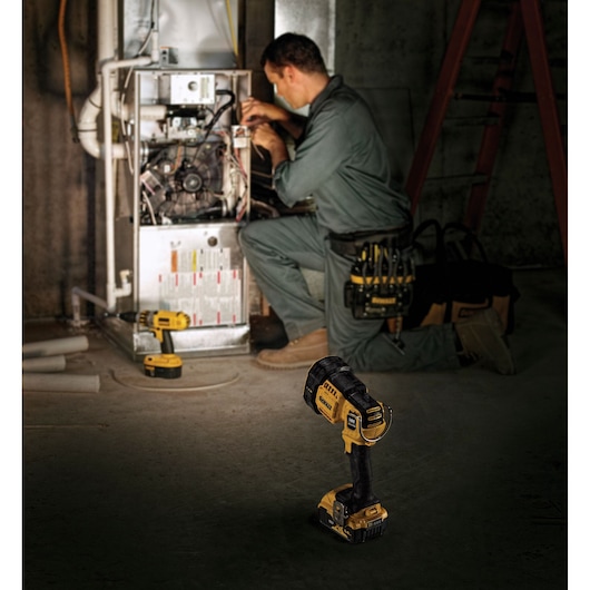 A DEWALT DCL043 cordless handheld spotlight is placed on the floor in the foreground, while a worker in coveralls is servicing an electrical unit in a dimly lit industrial setting.
