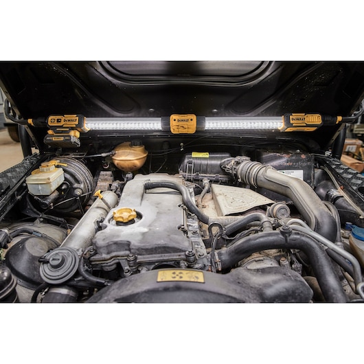A DEWALT DCL045 LED work light is mounted under the open hood of a vehicle, illuminating the engine compartment for better visibility while working.