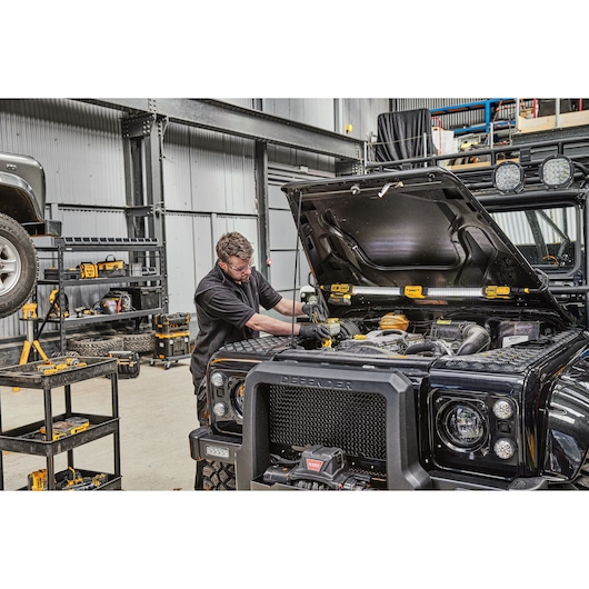 A person is working on the engine of a black Land Rover Defender inside a workshop. There are DEWALT tools visible on the shelves and next to the vehicle, and the DEWALT DCL045 work light is positioned under the open hood to illuminate the engine area.