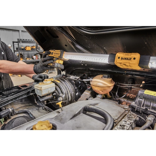 A mechanic uses a DEWALT DCL045 work light to illuminate the engine bay of a vehicle. The work light is mounted under the raised hood to provide bright lighting for maintenance and repair tasks.