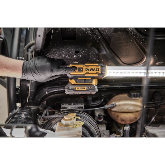 A gloved hand is holding a DEWALT DCL045 cordless LED work light under the hood of a vehicle, illuminating the engine area.