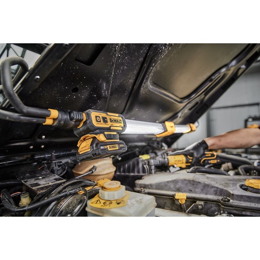 A DEWALT DCL045 LED work light mounted under a car hood, illuminating the engine bay while a worker uses a tool for repairs.