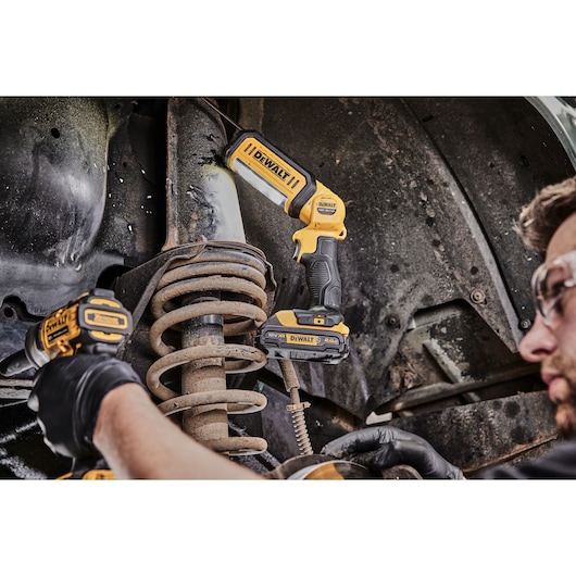 A DEWALT DCL050 LED work light is being used to illuminate an automotive suspension system while a person wearing safety glasses and gloves works with another DEWALT tool.