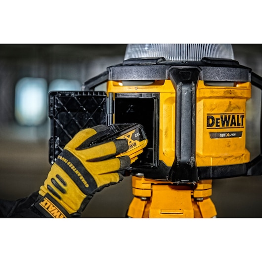 Gloved hand inserts battery into a yellow DEWALT work light. Close-up view of battery compartment and tool.