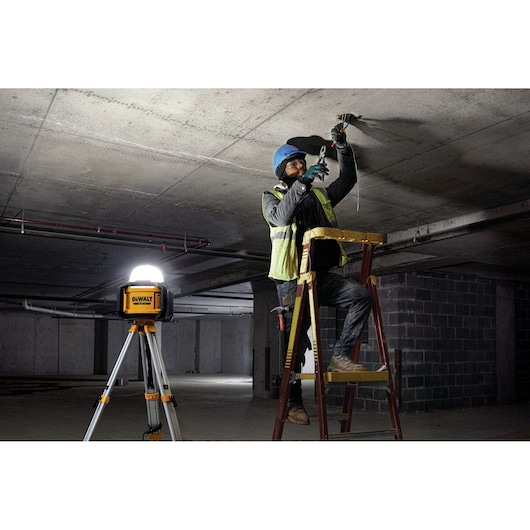 DEWALT DCL074 tripod work light illuminates a worker installing wiring on a ladder in a dimly lit construction site.