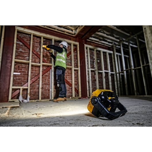 DEWALT DCL077 work light on floor in a construction site, worker in background using tools near wooden framing.