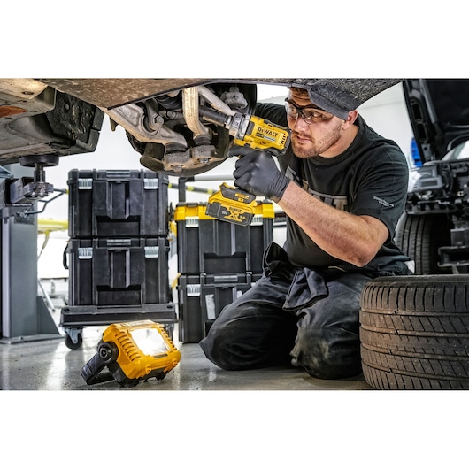 Mechanic uses DEWALT tools and LED work light under a car, with tool storage boxes visible in the background.
