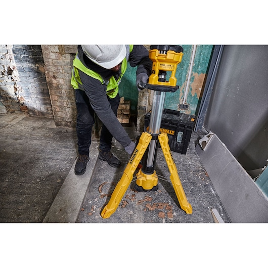 Worker adjusts DEWALT DCL079 yellow LED tripod light in a construction site with tools and concrete debris visible.